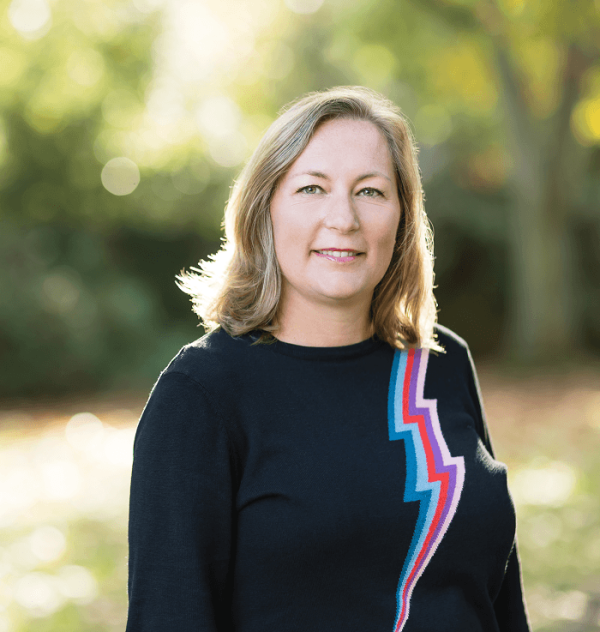 Sonia is standing in front blurred trees smiling wearing a black jumper
